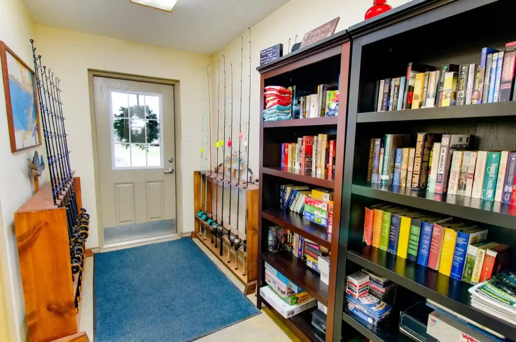 A small room features a bookshelf filled with books and board games on the right, with folded clothes on the top shelf. Fishing rods are neatly organized on the left side. The back door with glass panes opens to an outdoor view. A blue rug covers the floor.