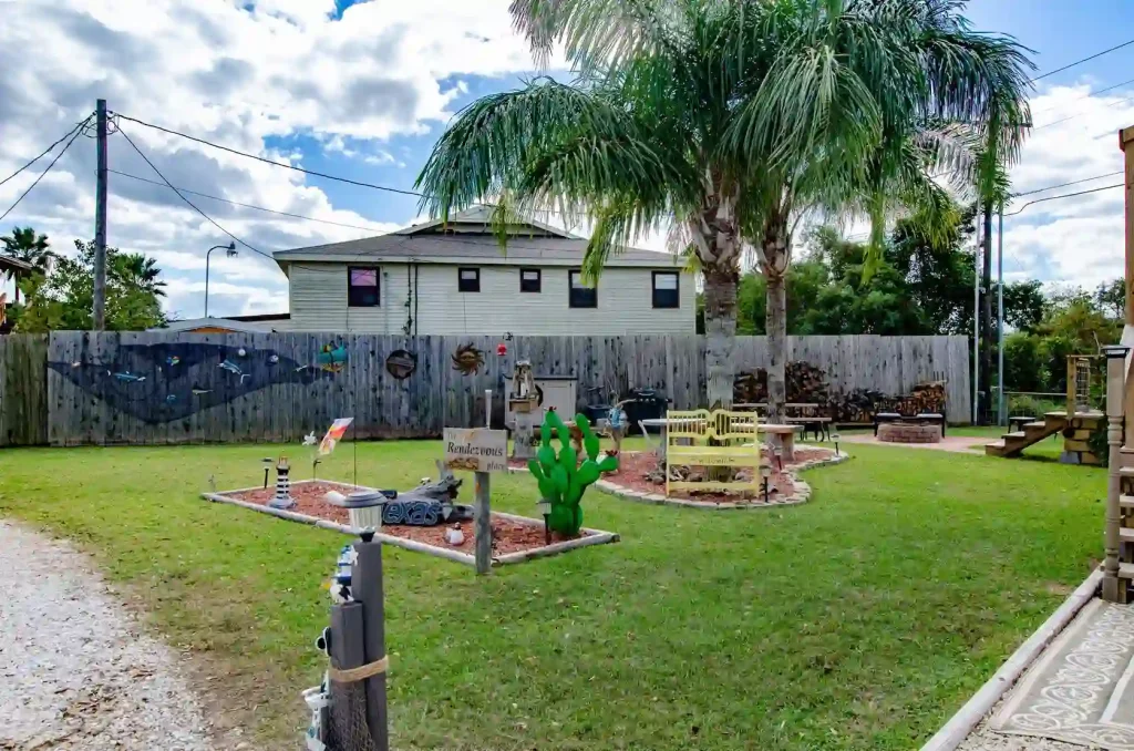 A vibrant garden with playful decorations, including a green cactus sculpture, colorful birdhouses, and quirky outdoor art. A yellow bench sits under tall palm trees. A wooden fence and a house are visible in the background under a partly cloudy sky.