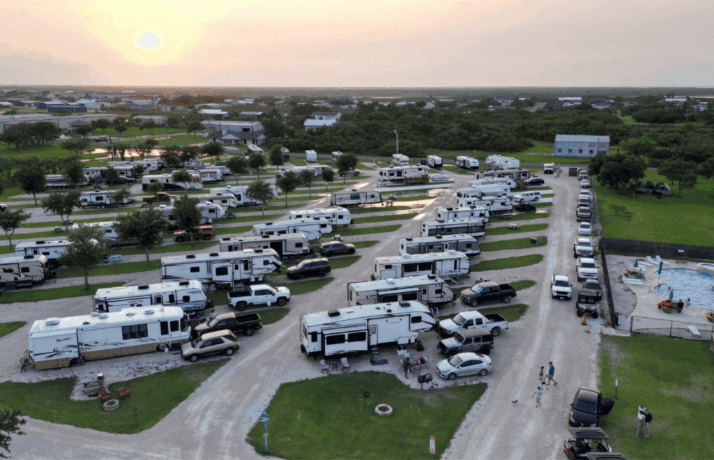 Aerial view of a busy RV park at sunset. Numerous RVs are parked in rows, with several vehicles next to them. There is a pool area with people gathered nearby. The surrounding area is green with trees and buildings in the distance. The sky is tinged with the soft colors of sunset.